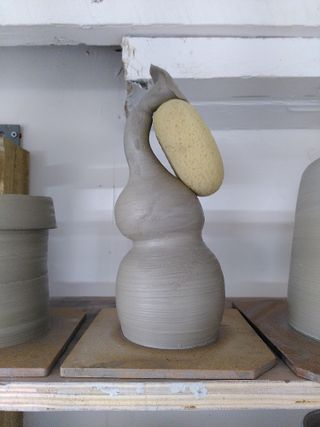 Close up of a clay vessel drying on a shelf: a double gourd form shaped as an elephant throwing water with its trunk, with a sponge supporting the trunk