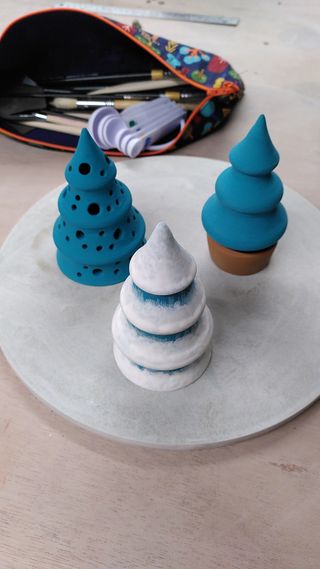 Three ceramic pine trees coated in blue gree underglaze, one of them with an extra coat of white resembling snow and one of them with a brown pot at the bottom.
