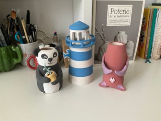 Painted ceramics: a panda vase holding a flower pot with a sprouting plant, a lighthouse stripped in blue, a brown bat with purple wings
