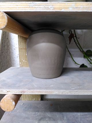 Trimmed and burnished clay form waiting to dry on a shelf. The form angles up outwards before rounding to a flat top with a hole.