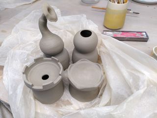 4 leather hard clay forms on a plastic sheet: a tower, a circus tent, a deformed double gourd and a head-and-body vase