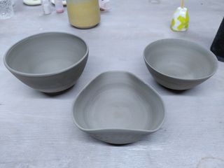 Three stoneware thrown bowls, leather hard and their form clean after trimming. The central one has its shape altered by flattening its wall to form a triangle.
