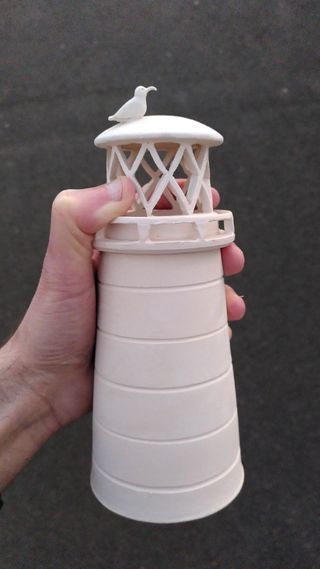 A hand holding a bisque-fired-white ceramic lighthouse with diamond carved light openings and a seagul with a delicate beak on the roof.
