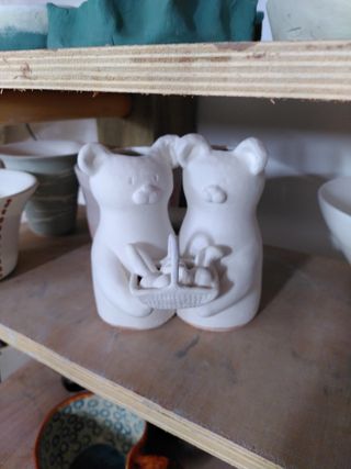 A ceramic piece on a shelf covered in a coat of white glaze: two bears holding each other by the shoulder while holding a bread basket.
