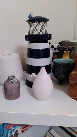 Picture of ceramic wares on a shelf, focused on a tiny bulbous bud vase with a mouse on its collar and a blue and white striped lighthouse with a seagull on its roof.