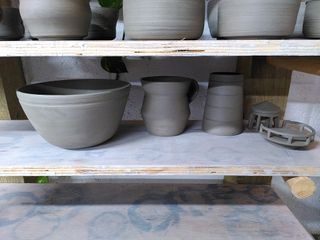 4 clay pieces waiting to dry on a shelf: a curvy clay bowl (with a carved stripe towards the rim), an angular clay mug  and the body, top and railing of a lighthouse.
