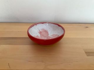 Little red bowl, on top of a raw ceramic foot, glazed white inside with thin blue splatters and red brush marks.