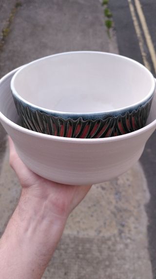 A hand holding two stacked clay bows, inner one is glazed blue with flaming carvings, outer one is white from its bisque firing.