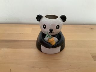 Little vase in the shape of a wide bellied panda holding a yellow pot where a green plant with two leaves is growing.

**Note** Outside glaze is very thin in places. Inside is good, though, and the vase is watertight.