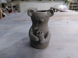 A little stoneware vase ready to dry before its bisque firing. It is shaped as a little bear holding a pot of which a plant is sprouting with 3 leaves along its stem.
