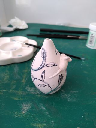 Back picture of a mini bud vase with a mouse poking its nose on the left of its collar, with marker drafting a vine-y pattern on the vase properly drawn in green underglaze.