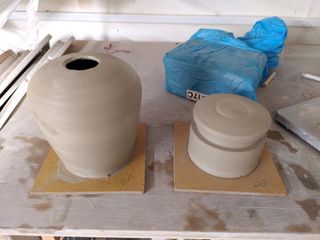 High angle shot of two clay forms on their bats after throwing: a table lamp base tapering at the bottom and curving inwards at the top and a closed cylinder with a large groove around the top.