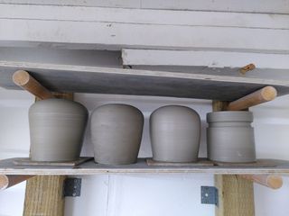 Clay forms drying on a shelf: 3 table lamp bases tapering at the bottom before curving inwards and a closed cylinder with a large groove at the top