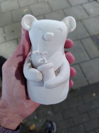 A bisque fired vase in the shape of a bear holding a pot with a growing plant.
