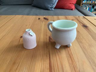 Two ceramic pieces painted in glaze. A tiny pink bottle vase, inside light blueish-green with a butterfly on the collar whose wings match the inside. A cauldron shape cup, light grey outside and light blueish-green inside overflowing at the rim in a thick layer.
